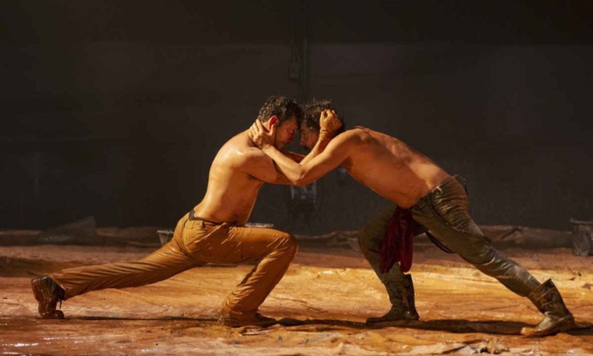 Atores sem camisa lutam e est&atilde;o de lado, em p&eacute;, sob ch&atilde;o de lama, em cena da pe&ccedil;a Tom na fazenda