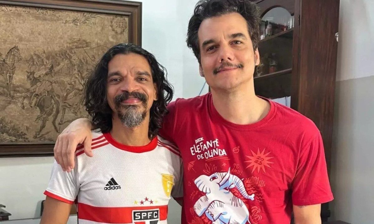 Atores Kaiony Ven&acirc;ncio e Wagner Moura est&atilde;o abra&ccedil;ados e olham sorrindo para a c&acirc;mera