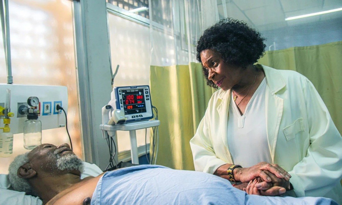 Atriz Zez&eacute; Motta, no papel de enfermeira Regina, est&aacute; no ambulat&oacute;rio, de jaleco branco, e segura a m&atilde;o de paciente idoso, deitado na cama, em cena da s&eacute;rie Invulner&aacute;veis