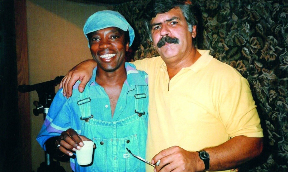 Milton Nascimento, usando bon&eacute; e camisa azuis, e Dori Caymmi, usando camisa amarela, est&atilde;o abra&ccedil;ados e sorriem para a c&acirc;mera em foto dos anos 1980
