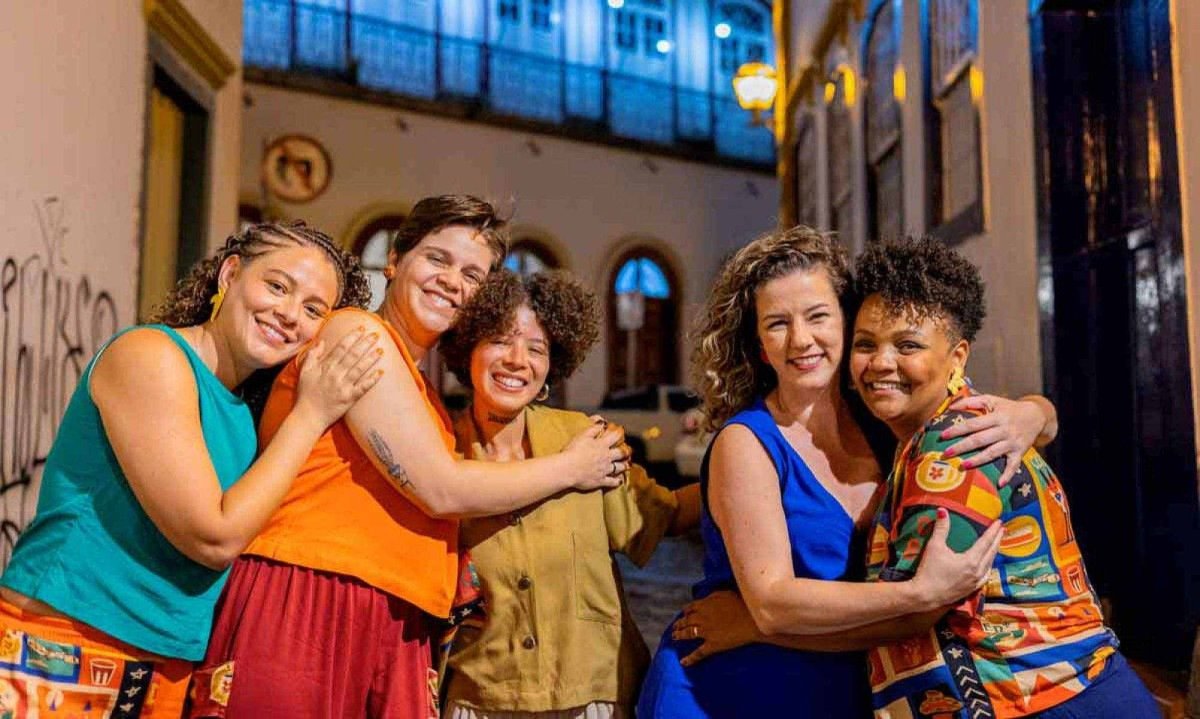 Cinco mulheres que integram o grupo musical Chorosas est&atilde;o abra&ccedil;adas, lado a lado, e sorriem para a c&acirc;mera