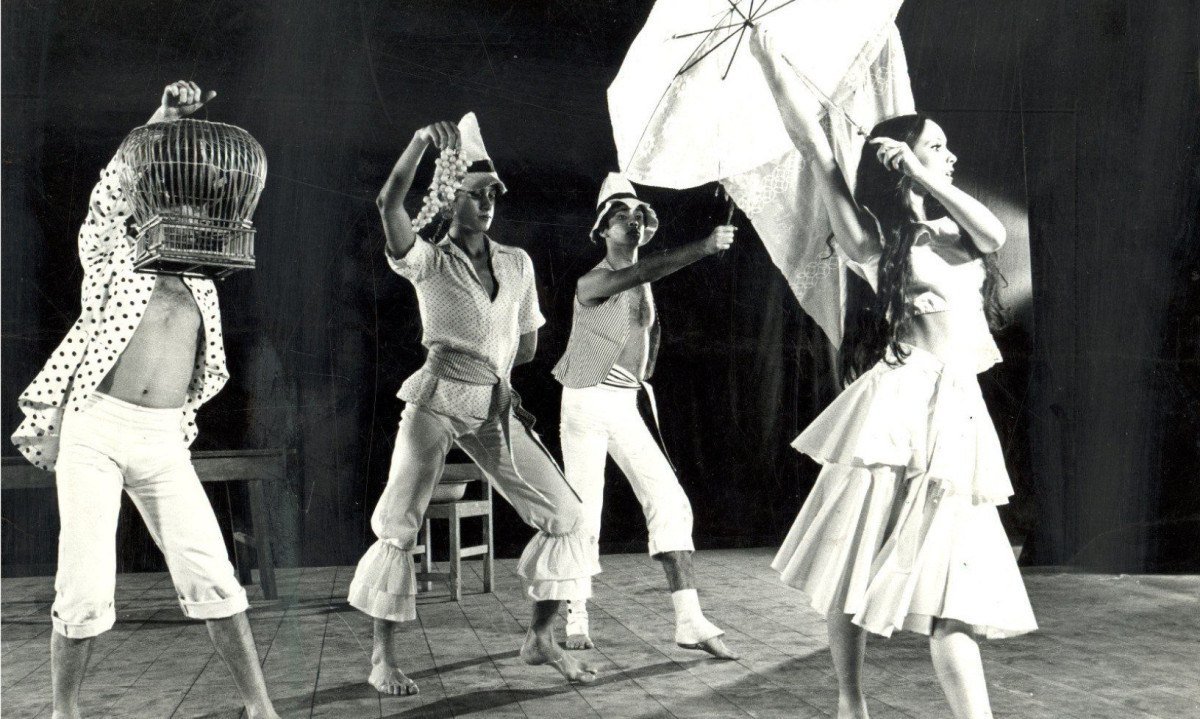 Bailarina segura guarda-chuva. Ela est&aacute; &agrave; frente de bailarino que segura gaiola e de outros dan&ccedil;arinos em cena de Maria Maria, espet&aacute;culo do Grupo Corpoi