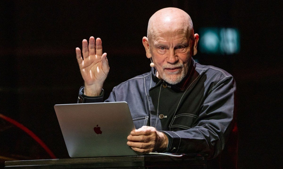Ator John Malkovich est&aacute; diante de laptop e levanta a m&atilde;o direita em cena da pe&ccedil;a The infamous Ram&iacute;rez Hoffman