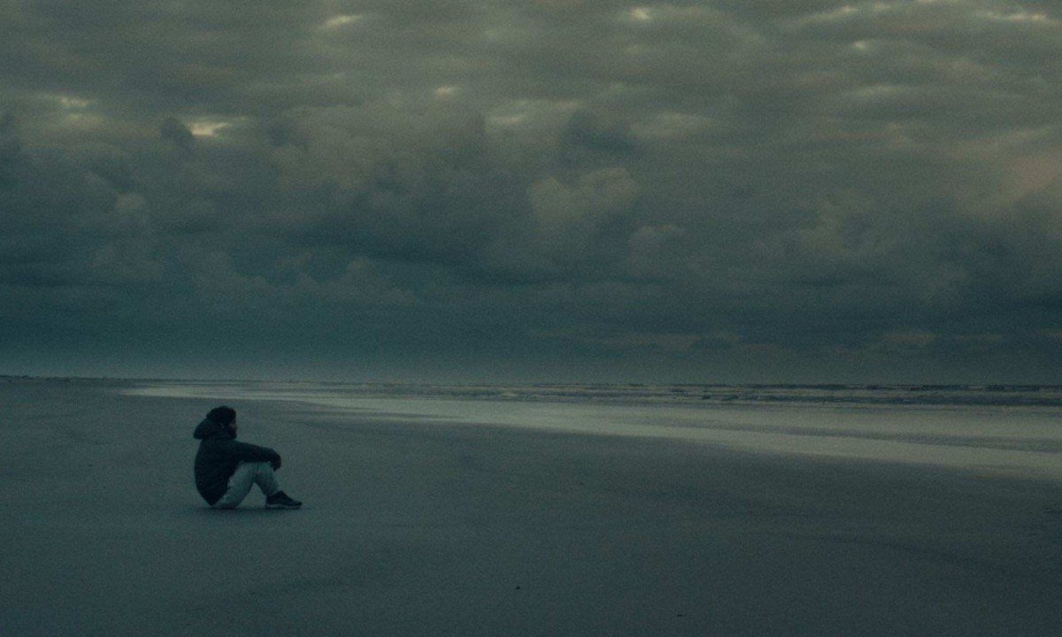 Ator Gabriel Leone est&aacute; sentado na areia, observando o mar, em cena do filme Barba ensopada de sangue