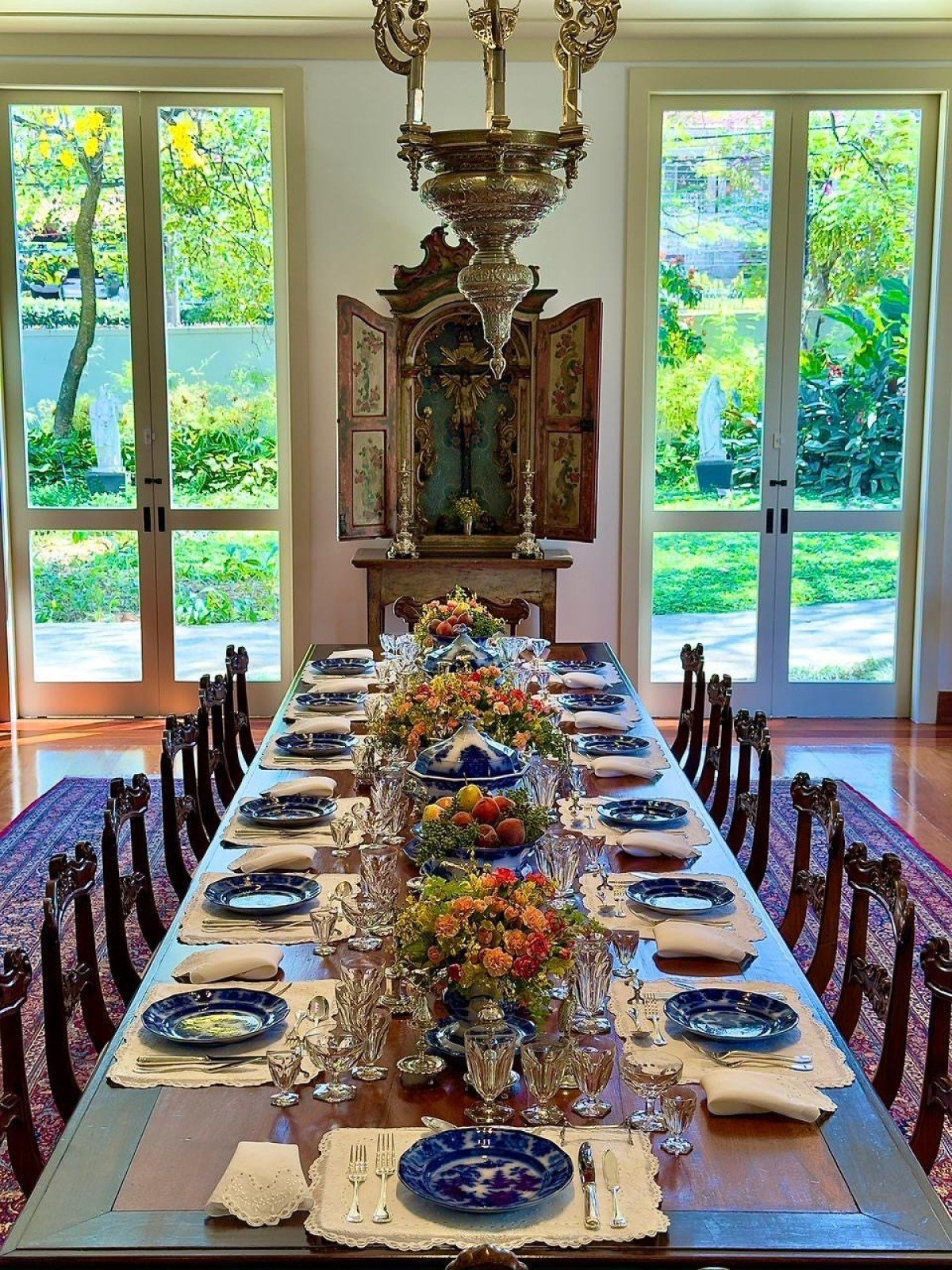 Mesa posta em sala de jantar com pratos talheres flores toalha com lustre e portas de vidro ao fundo
