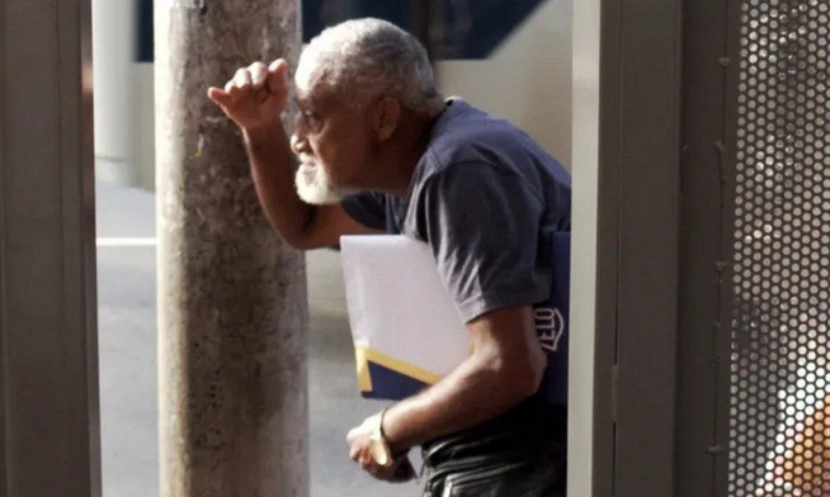 Senhor est&aacute; de lado na rua, protegendo os olhos do sol com a m&atilde;o e carregando pasta de documentos debaixo do bra&ccedil;o em fotografia feita pelo rapper Materia Prima