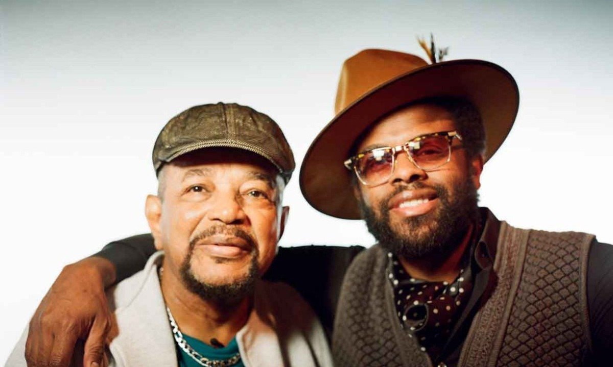 M&uacute;sicos Carlos Daf&eacute; e Adrian Younge est&atilde;o abra&ccedil;ados e sorriem para a c&acirc;mera. Ambos usam chap&eacute;u