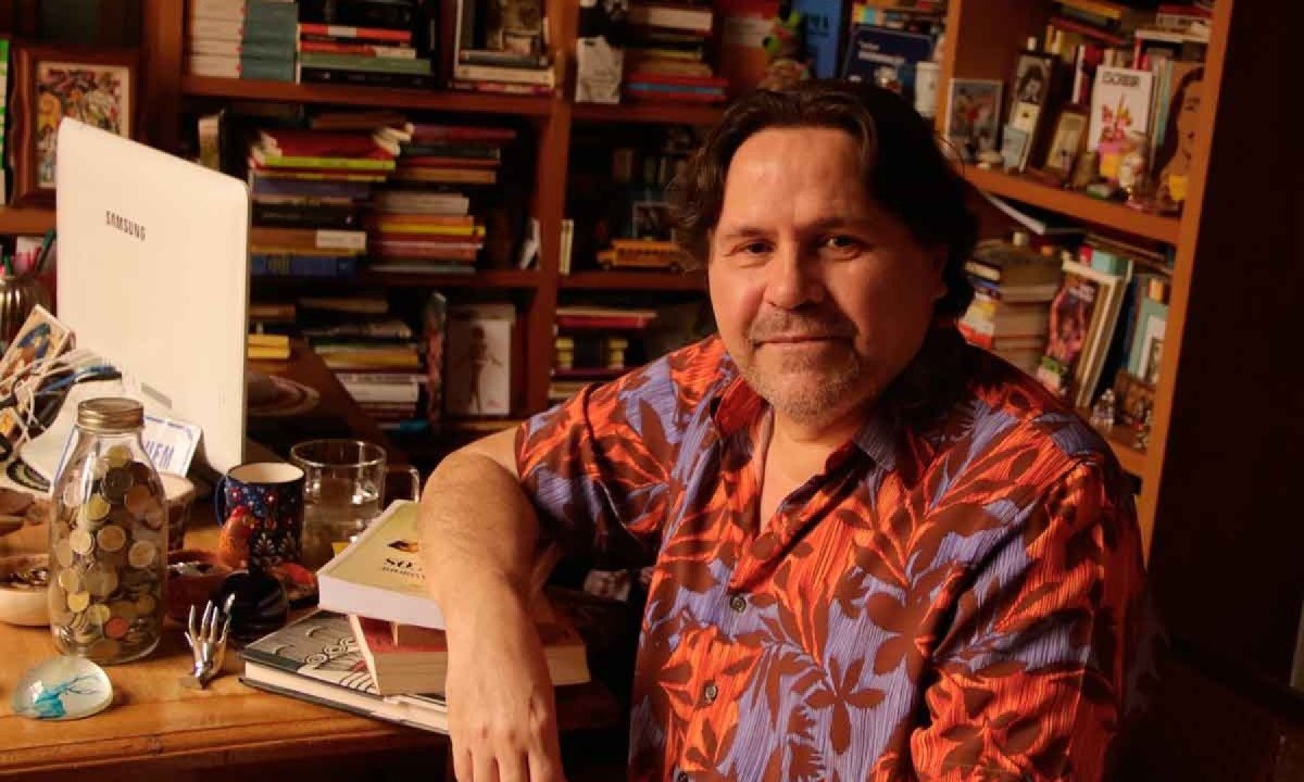 O escritor Marcelino Freire usa camisa estampada, est&aacute; de lado e olha para a c&acirc;mera. Atr&aacute;s dele se v&ecirc; estante lotada de livros