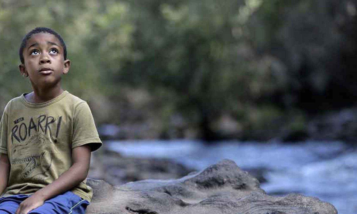 Menino sentado em pedra, &agrave; beira de rio, olha para cima em cena do filme A mensagem de Jequi