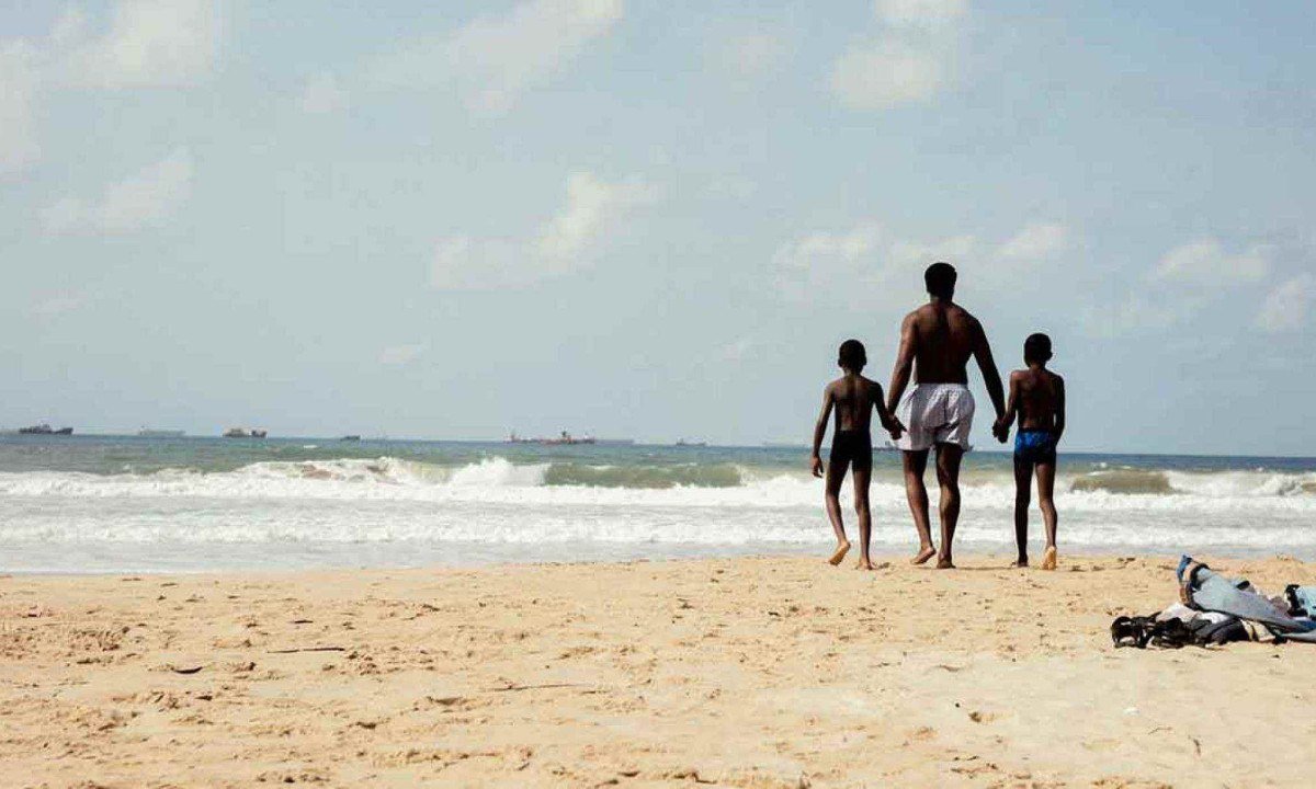 Dois meninos est&atilde;o de m&atilde;os dadas com o pai em frente ao mar na cidade de Lagos, em cena do filme A sombra do meu pai