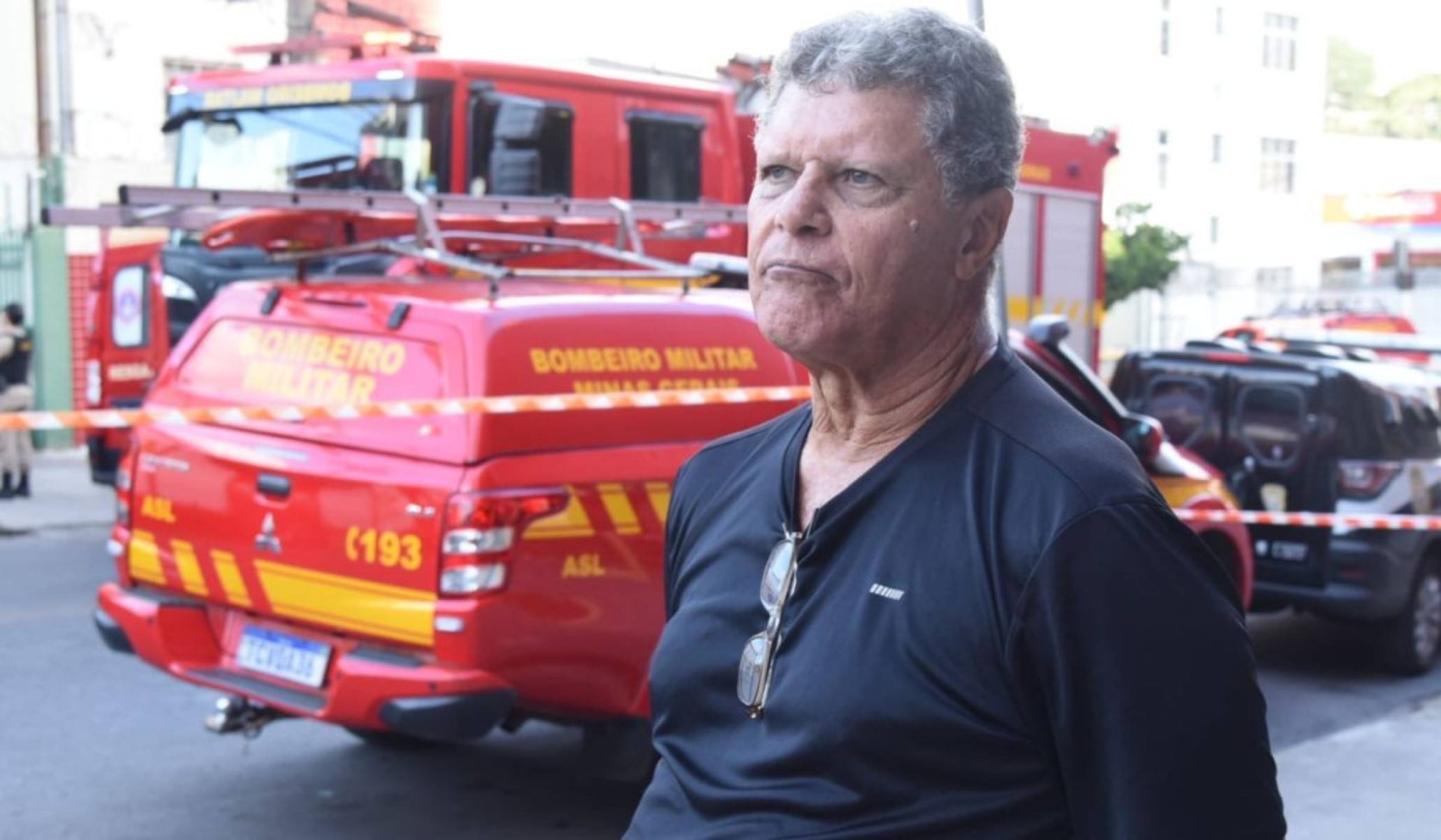 Argeu &eacute; um homem branco, que veste camisa preta e tem cabelo grisalho. Ele esta no centro da imagem. Ao fundo, um carro vermelho com detalhes amarelos dos bombeiros 