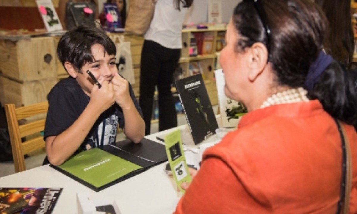 Cac&aacute; Gontijo, de 11 anos, est&aacute; sentado em mesa de evento liter&aacute;rio e autografa livro para senhora sentada &agrave; sua frente