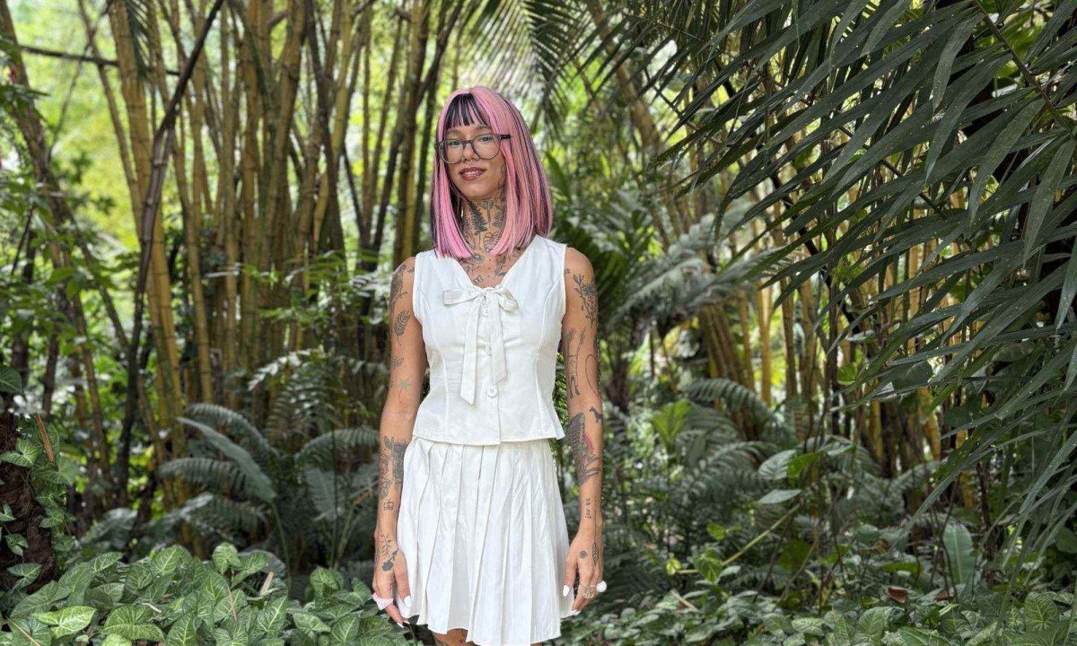 Artista visual Efe Godoy olha para a c&acirc;mera e posa em jardim repleto de folhagens e &aacute;rvores. Ela usa &oacute;culos, vestido branco e cabelos cor-de-rosa