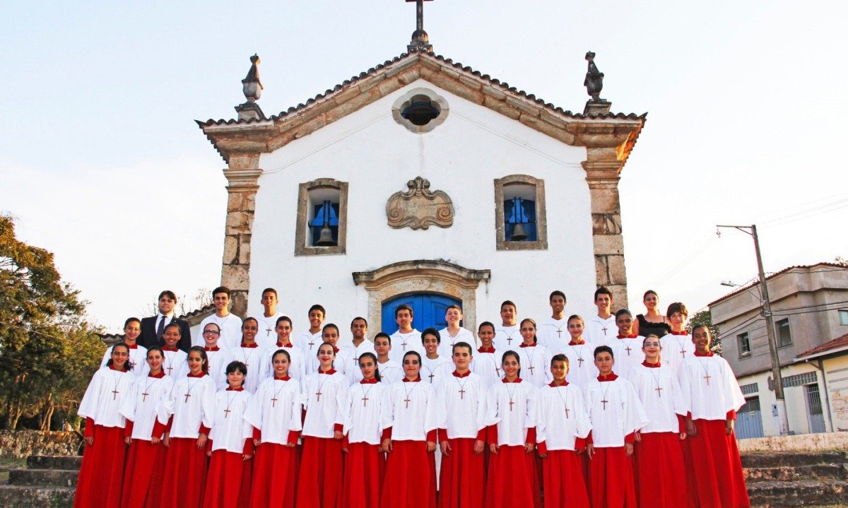 Cantores do Coral Canarinhos de Itabirito vestem figurino branco e vermelho e posam em frente a igreja antiga