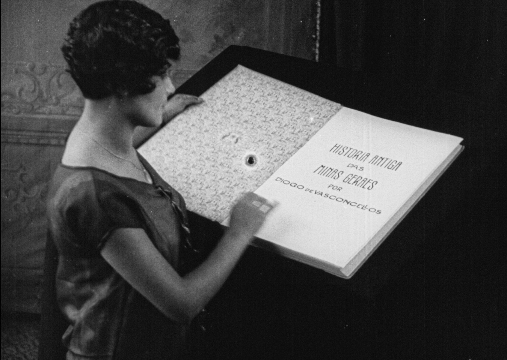  Em cena de filme de 1926, mulher abre imenso livro de Hist&oacute;ria de Minas Gerais escrito por Diogo de Vasconcellos