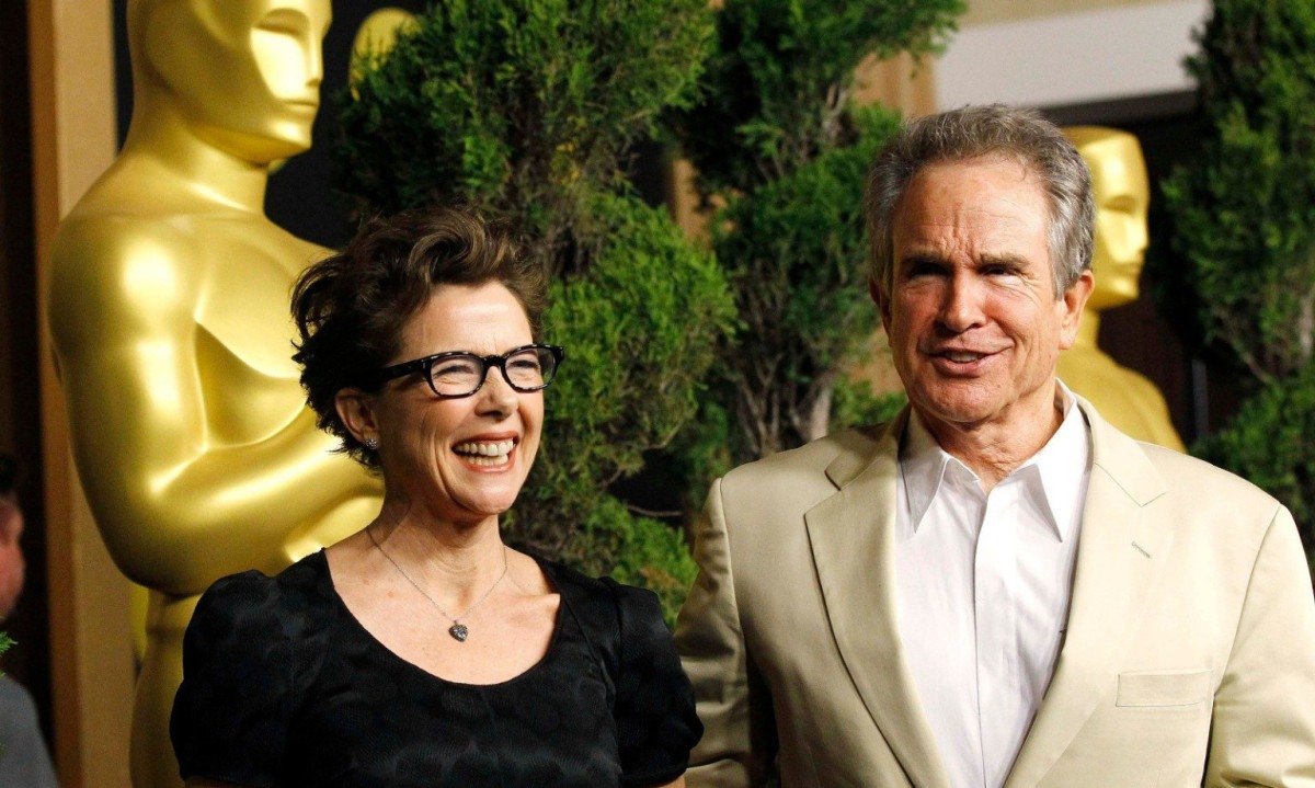 Na festa do Oscar em 2011, atores Annette Bening e Warren Beatty sorriem para os fot&oacute;grafos. Atr&aacute;s deles h&aacute; imensa est&aacute;tua dourada do Oscar