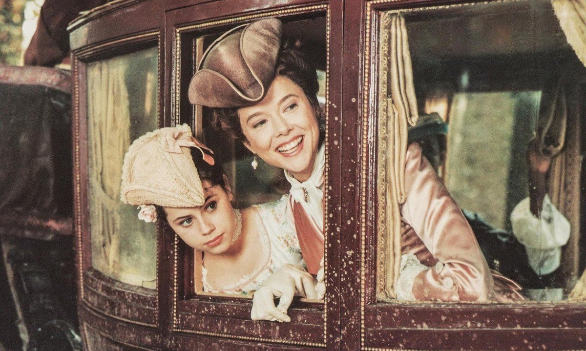 Atrizes Fairuza Balk e Annette Bening sorriem na janela de carruagem, vestindo roupas antigas, em cena do filme Valmont: Uma hist&oacute;ria de sedu&ccedil;&otilde;es