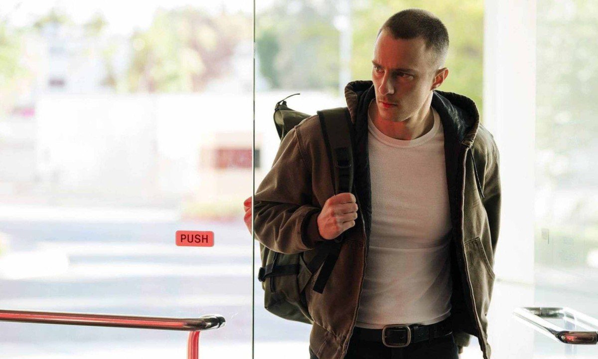 Drew Starkey abre porta de vidro em cena da s&eacute;rie Lucky. Ator veste casaco de couro e carrega mochila nas costas