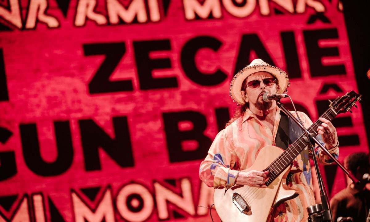 Geraldo Azevedo canta ao microfone enquanto atr&aacute;s dele letra da m&uacute;sica Mon ami, em dialeto angolano, &eacute; exibida em tel&atilde;o