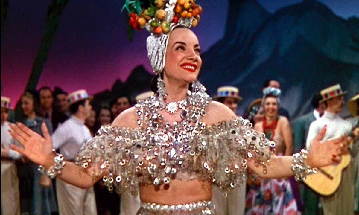 Carmen Miranda, vestida como baiana, abre os bra&ccedil;os, sorri e olha para o lado