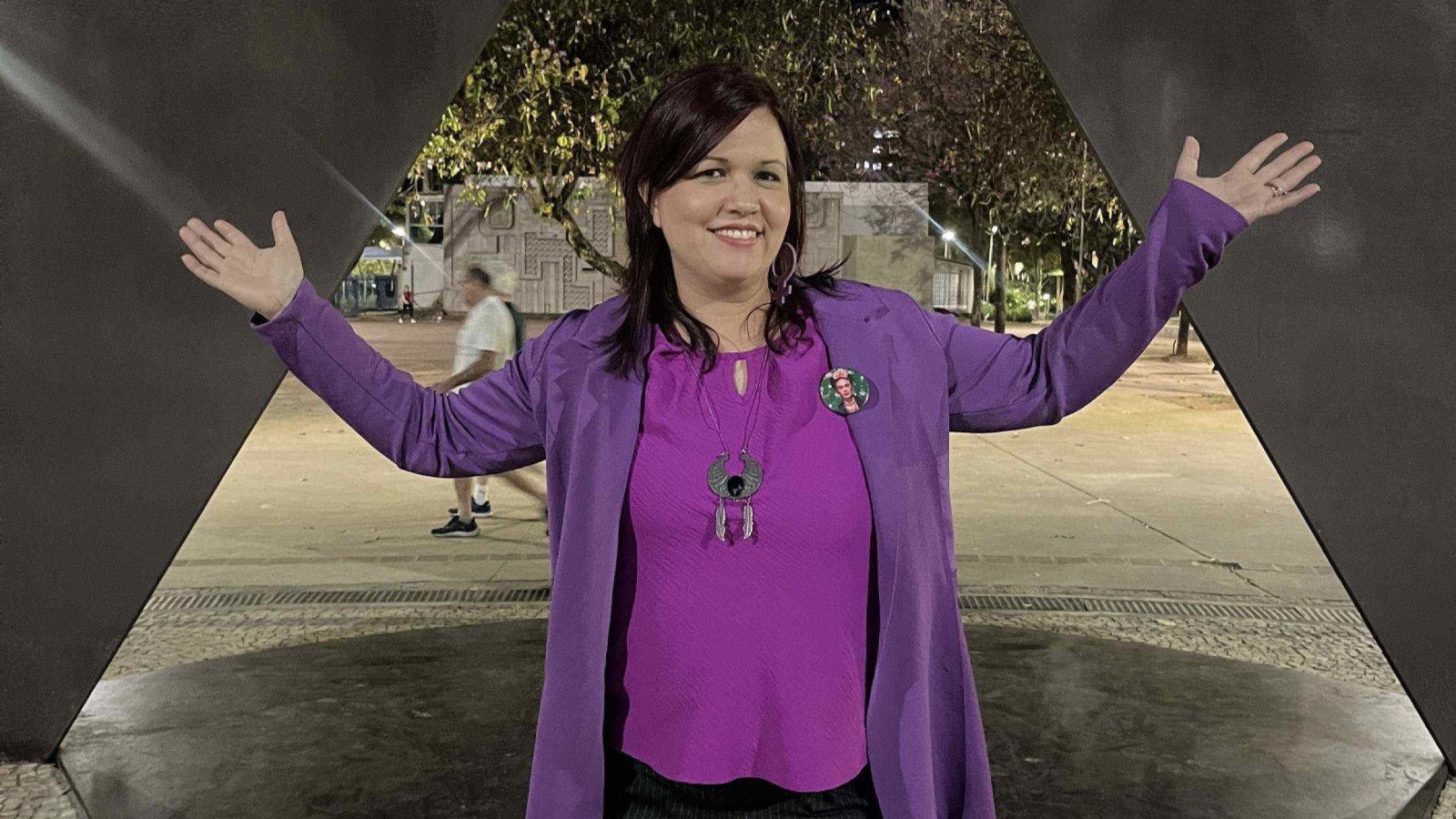   Escritora Brenda Pena usa blusa roxa, tem os bra&ccedil;os abertos diante de escultura de ferro e sorri para a c&acirc;mera