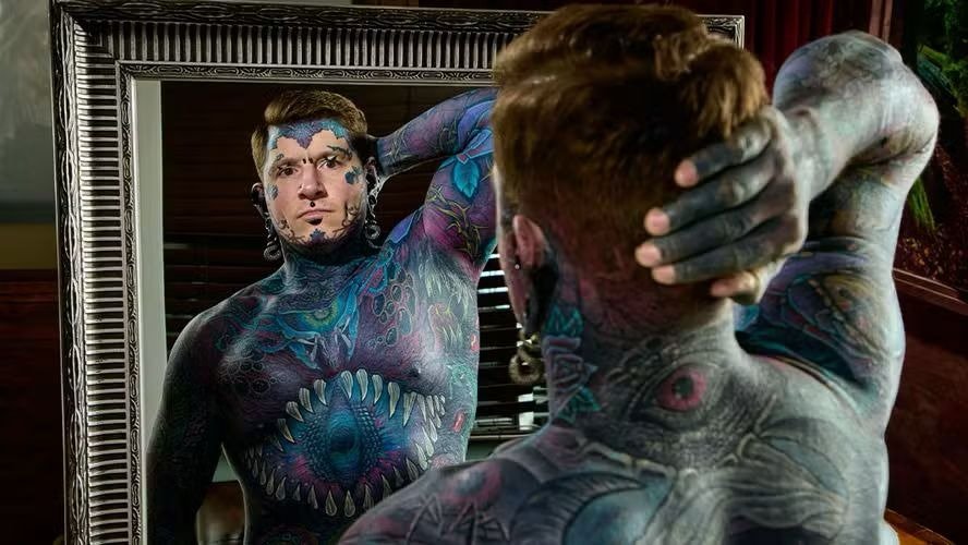 O canadense Ephemeral Remy, recordista mundial por suas m&uacute;ltiplas camadas de tatuagens, observa a arte que cobre seu corpo no espelho. -  (crédito: Guinness World Records)