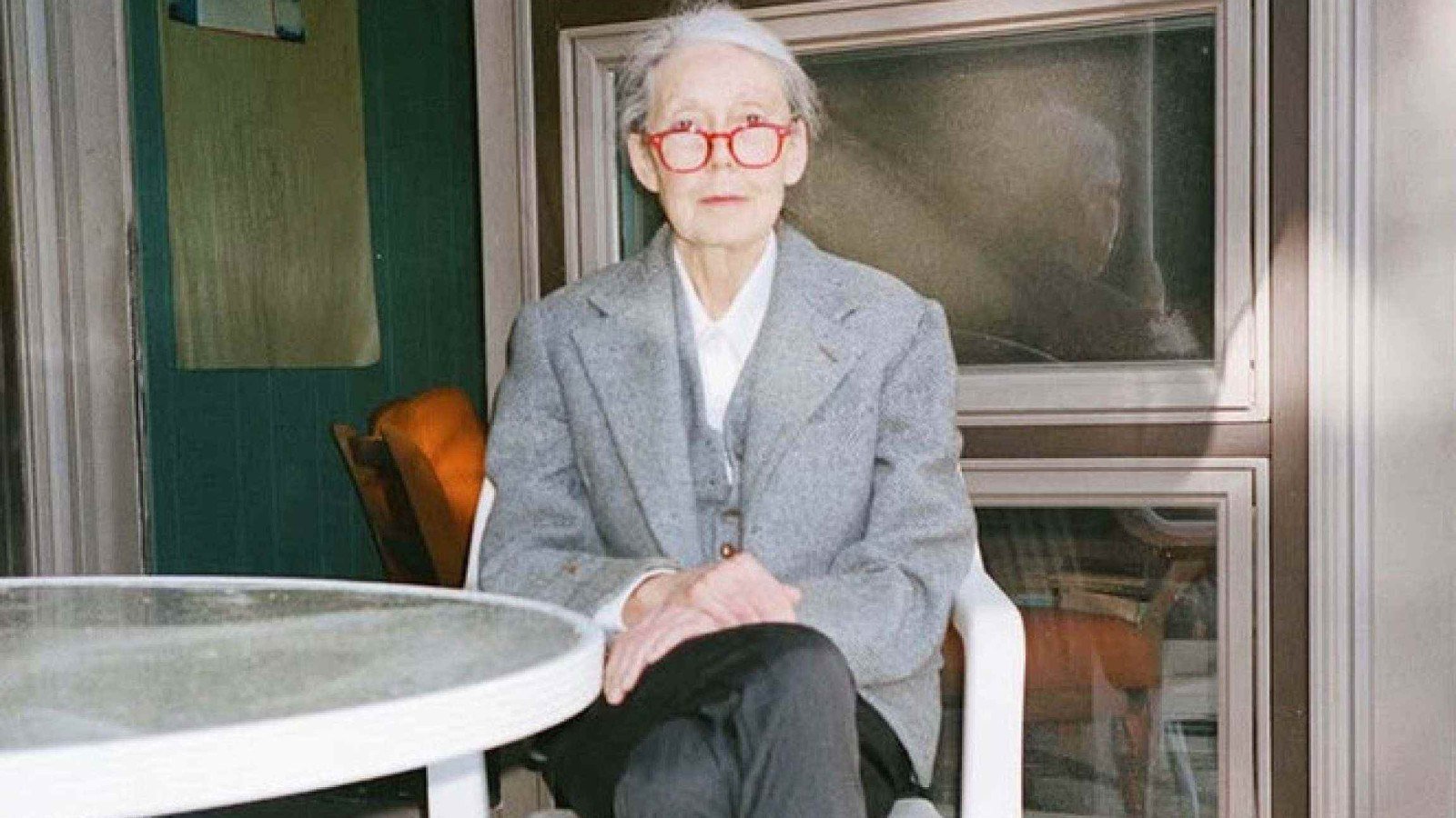 Escritora Anne Carson est&aacute; sentada, com as pernas cruzadas, &agrave; frente de mesa branca, e olha para a c&acirc;mera