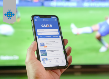 Uma pessoa segura um smartphone preto, com o aplicativo da Caixa Econômica Federal aberto na tela, em frente a uma televisão que exibe um jogo de futebol, simbolizando a nova casa de apostas da Caixa.  -  (crédito: Reprodução Educando seu bolso | Imagem gerada por IA)